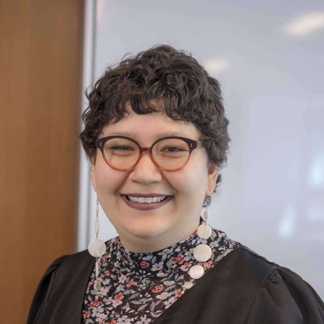 Portrait of Amy Pachai, a smiling woman with glasses and short curly hair wearing a brown top.