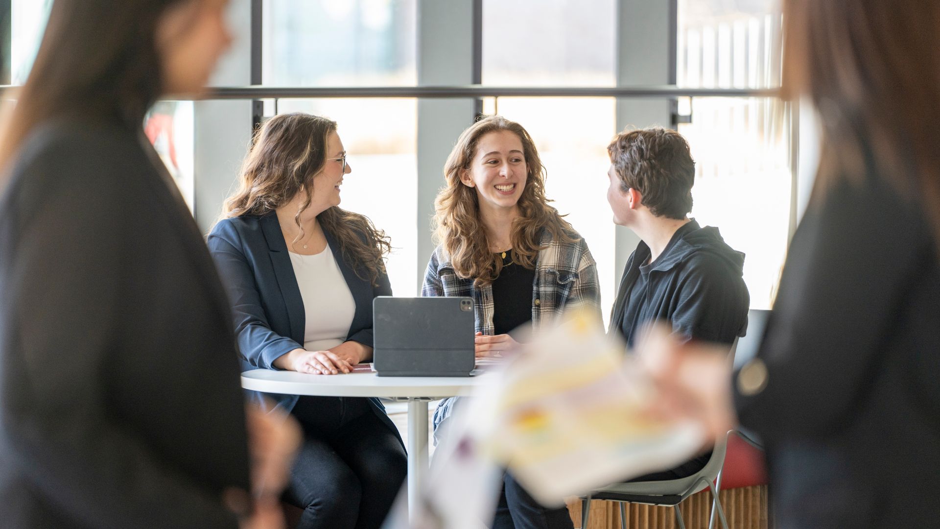Three DeGroote students are seated at a table around a tablet, talking and smiling.