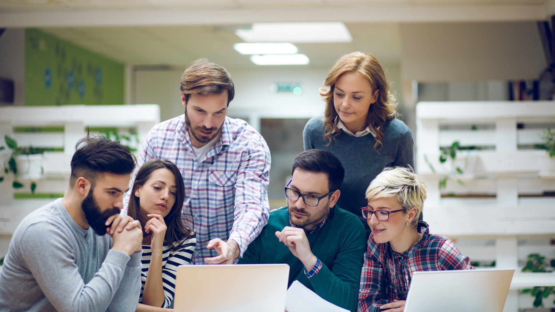 Six professionals huddled over a laptop, pondering, discussing, and pointing to the screen.