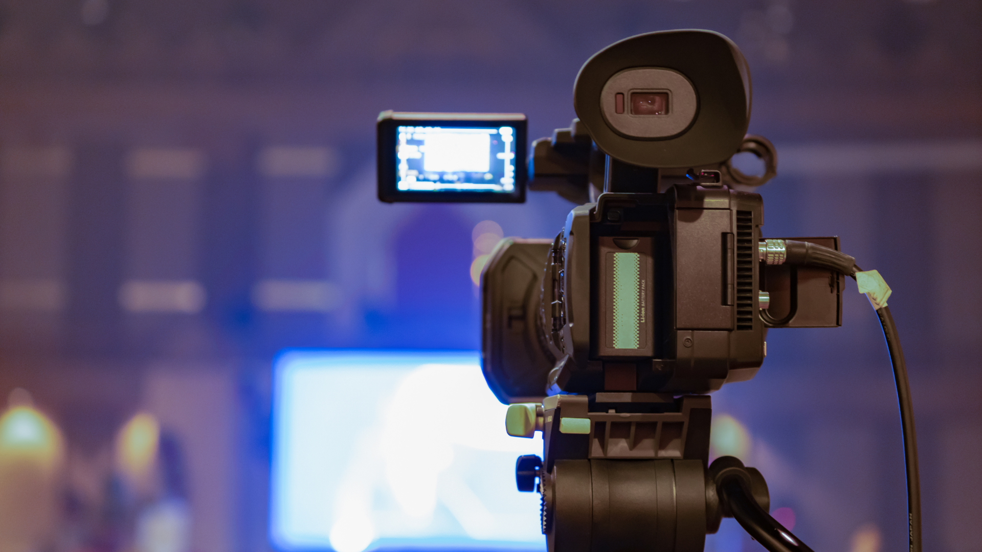 A professional video camera is set up indoors, focused on a blurred screen in the background.
