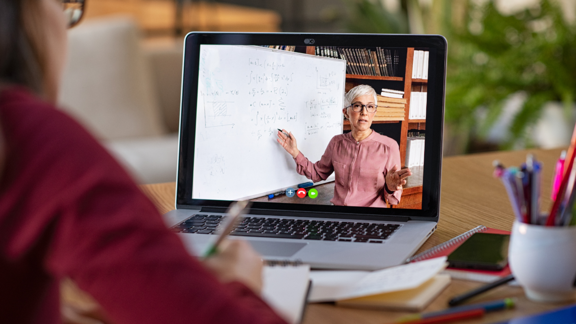 A female professor teaching remotely on the computer via a laptop screen, while a student is watching and writing on a table