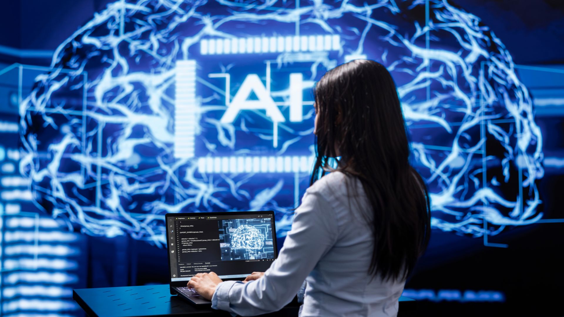 A person is working on a laptop with code displayed on the screen. In the background, there is a large digital representation of a brain with