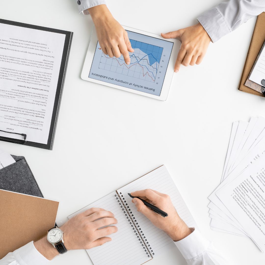 Two people work at a table. One points at a tablet showing a graph. The other writes in a notebook. Documents are scattered around.