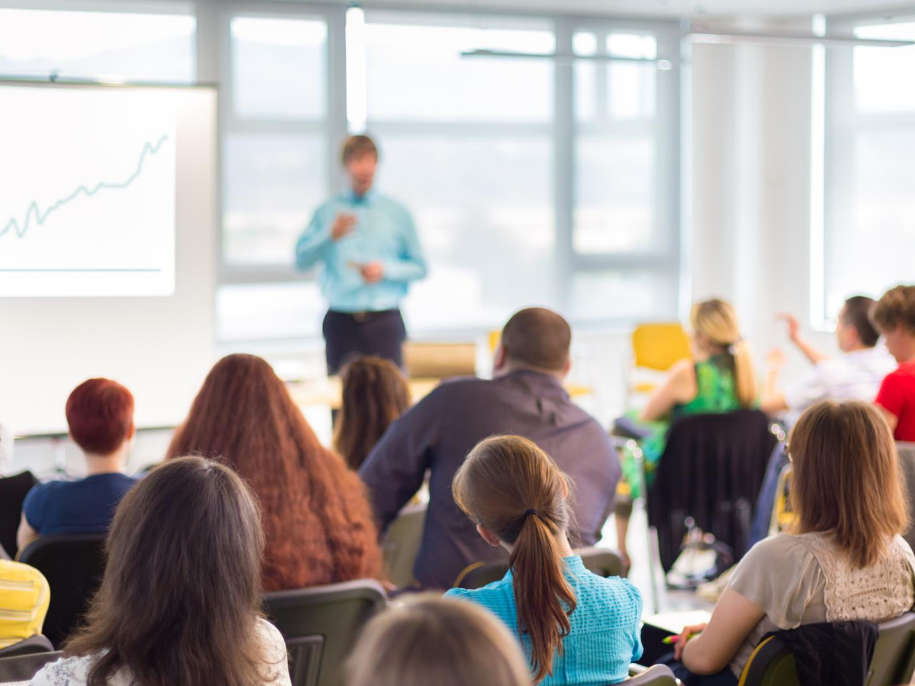 A crowd of people seated, listening to a speaker next to a projector screen.