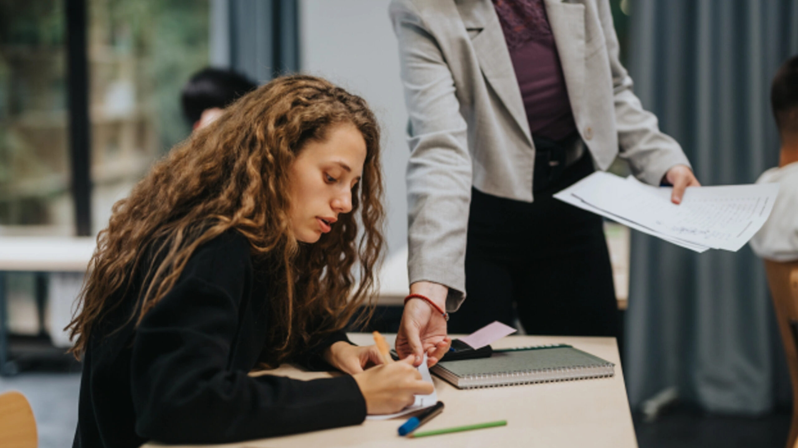 A student is writing on a paper while her professor gives guidance while pointing at specific part.