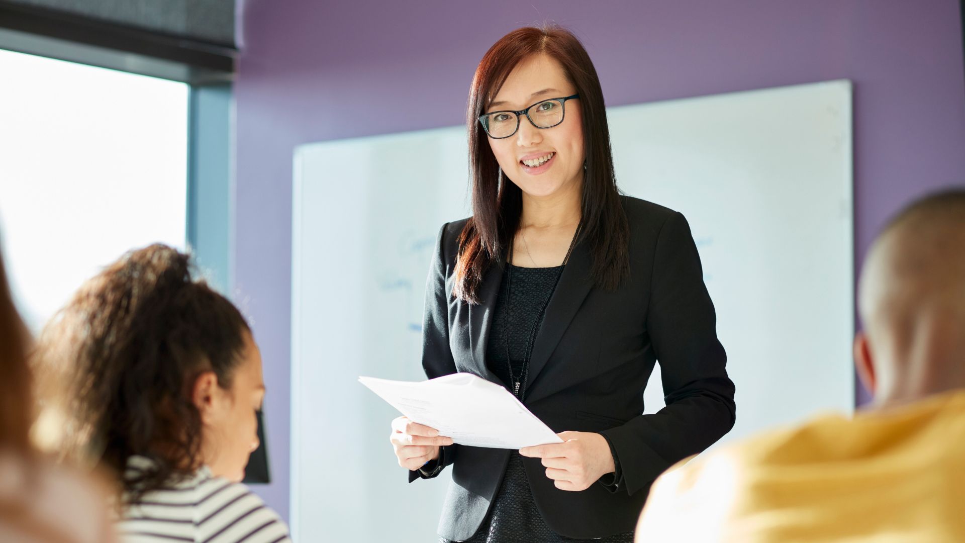 A woman in a black blazer is holding papers and speaking in front of a group, with a whiteboard in the background.