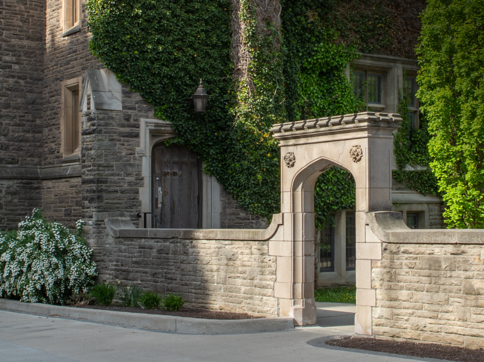 A stone archway entrance to a building. The building has a classic architectural style with stone walls and arched windows. Greenery, including bushes and trees, surrounds the entrance, adding to the serene and historic atmosphere of the scene