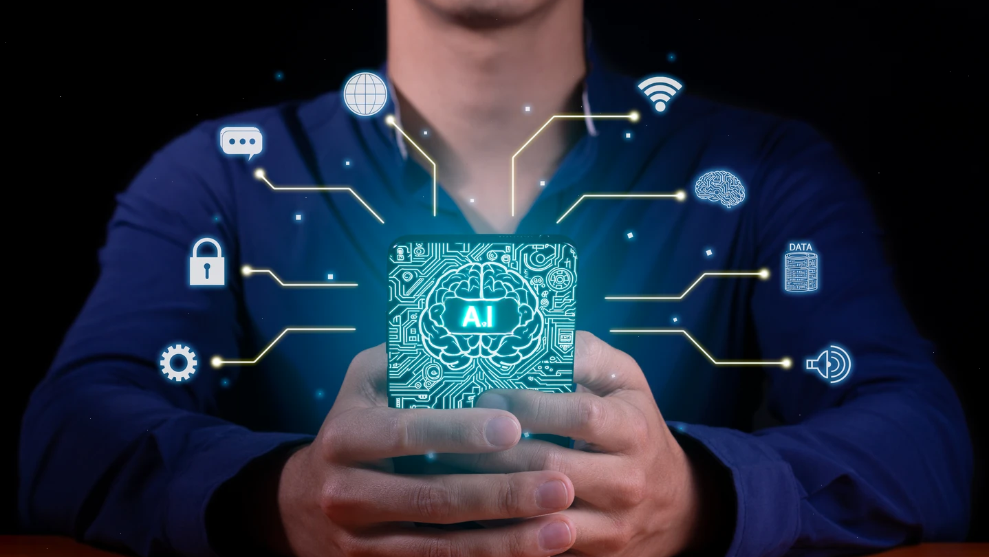 A person is holding a glowing cube with the letters 'AI' and a brain icon on it. The cube has lines extending outward, connecting to various icons representing technology and communication, such as a globe, Wi-Fi signal, lock, chat bubble, and data storage. This image visually represents the concept of artificial intelligence (AI) being central to various technological advancements and applications.