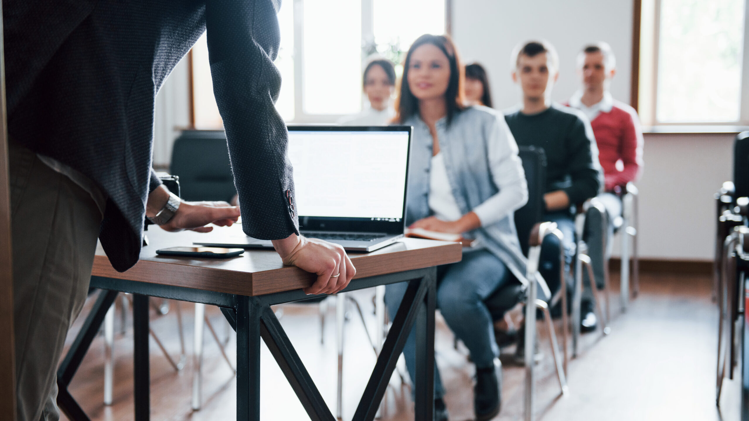 A group of people is seated in a classroom or meeting room, facing a person standing at the front. The person at the front is leaning on a table with a laptop and some papers. The room has large windows allowing natural light to enter. This image depicts an educational or professional setting, highlighting interaction and engagement among participants.