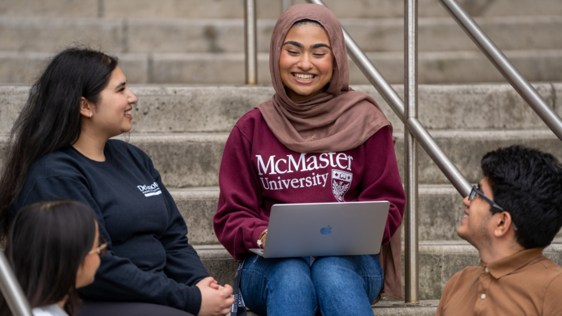 Four people are sitting on outdoor steps. One person is wearing a maroon 'McMaster University' sweatshirt and holding a laptop. The other three individuals are engaged in conversation with this person.