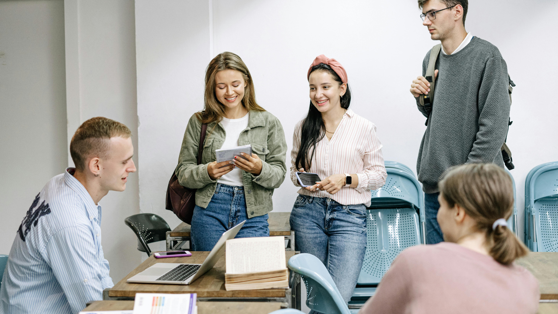 A group of five people sitting and standing around a table, studying together with books, notebooks, and a laptop, engaged in discussion.