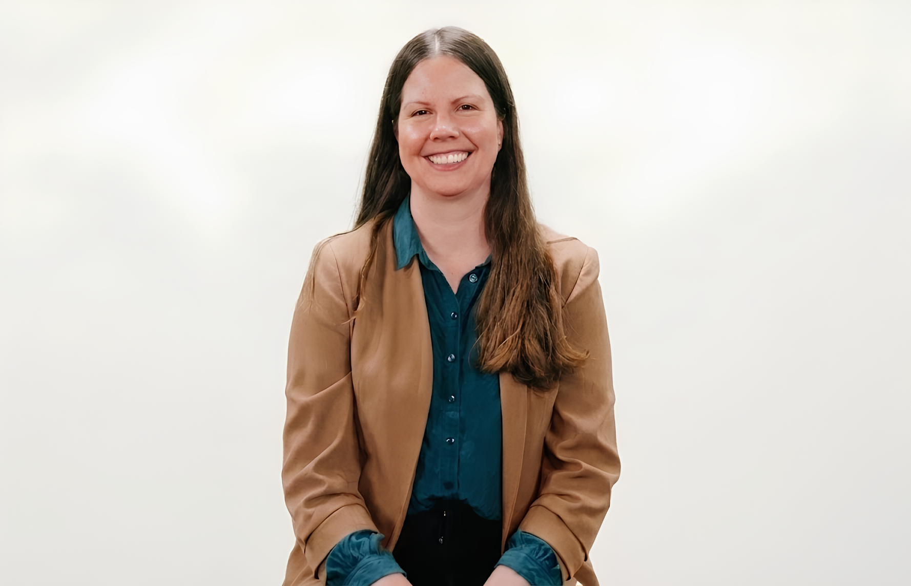A woman is smiling for a studio portrait while wearing a green collared shirt and a brown blazer.